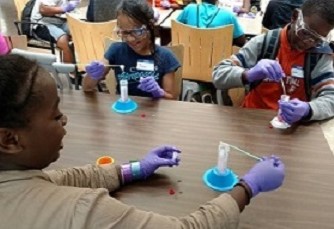 Three kids, sitting at a table wearing gloves and doing an experiment